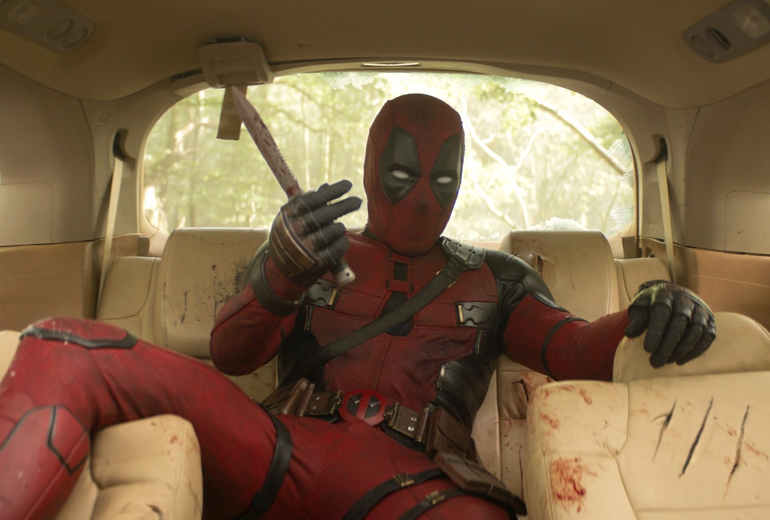 Deadpool (Ryan Reynolds) sprawls out, legs spread disconcertingly wide, in the back seat of a shredded, bloodied car in Deadpool & Wolverine